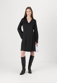 Čierne šaty s dlhými rukávmi a výstrihom do V, vyrobené z hladkej látky, vo voľnom strihu, s dĺžkou po stred stehien; v kombinácii s lesklými čiernymi čižmami po kolená.