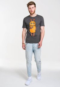 Camiseta gris con un personaje de dibujos animados de cuerpo naranja y detalles marrones, combinada con jeans azul claro y zapatillas blancas.