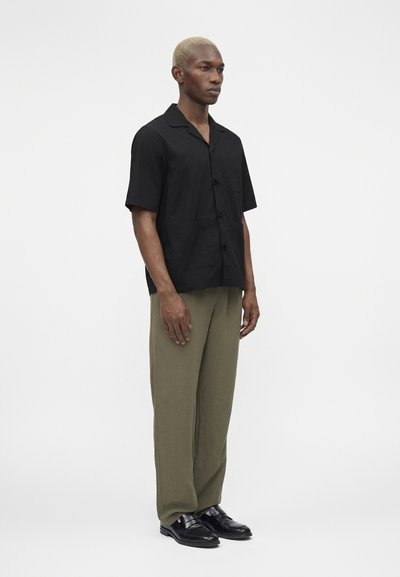 Άνδρας με κοντά ξανθά μαλλιά, που φορά μαύρο κοντομάνικο πουκάμισο, ελιάς πράσινο παντελόνι και μαύρα παπούτσια, στέκεται μπροστά σε μια απλή ανοιχτή φόντο.