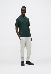 Camiseta polo verde oscuro combinada con pantalones jogger gris claro y zapatillas negras. La tela parece suave; el corte es relajado, sin patrones visibles.