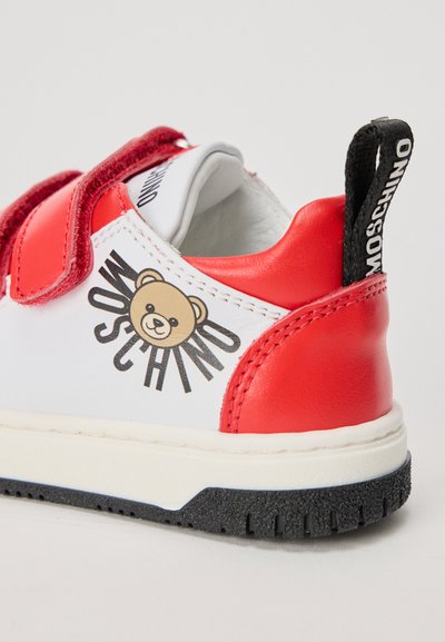 Červeno-biele tenisky s grafikou medvedíka a textom "Moschino". Uzavretie na suchý zips, mäkký kožený materiál a textúrovaná čierna gumová podrážka.