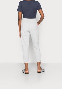 Osoba ubrana w jasnoszare przycięte spodnie z tylnymi kieszeniami, granatową koszulę z krótkim rękawem oraz szare sandały wsuwane, stojąca na białym tle.