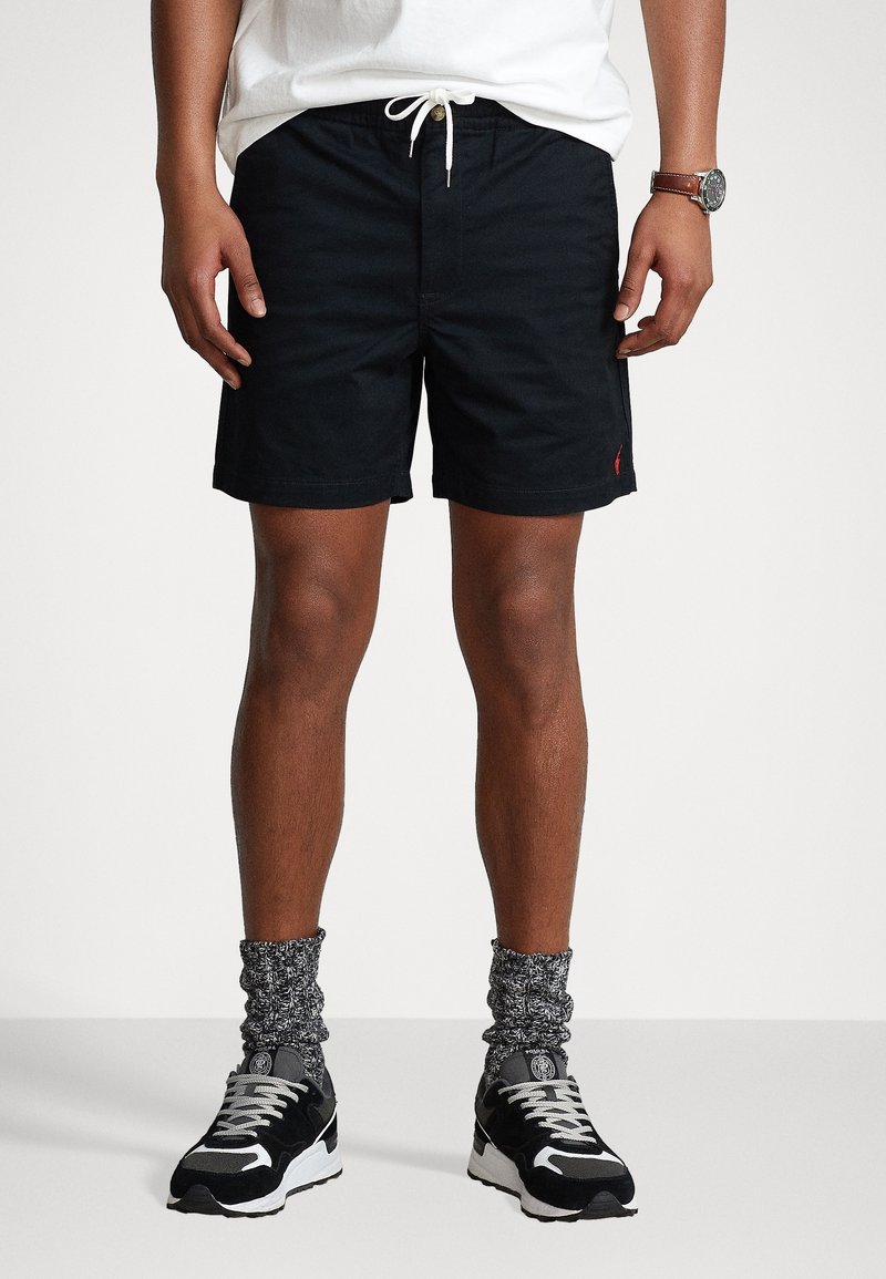 Man som bär svarta shorts med dragsnodd, vit skjorta, grå mönstrade strumpor, svarta och vita sneakers samt ett armbandsur, stående framför en vit bakgrund.
