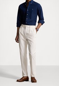 Marineblaues Hemd mit Knopfleiste, cremfarbene, gefaltete Chinos, braune Lederloafers. Slim Fit, glatte Textur, klassisches Design mit subtilen Markendetails.