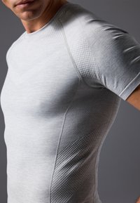 Světlé šedé sportovní tričko s přiléhavým designem, texturované panely na ramenou a hladká tkanina, která obepíná tělo.