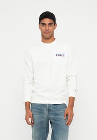 Muž v bílé mikině se nápisem "1985 Tommy Jeans" a modrých džínech, stojící proti hladkému bílému pozadí.