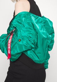 Groene bomberjas van glanzend materiaal, met elastische onderrand en een rode label met de tekst "VERWIJDER VOORDAT U VLIEDT." Heeft een vrouwtjeszak met rits.