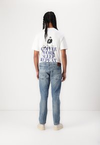 Weißes Baumwoll-T-Shirt mit blauer Grafikschrift "KAFFEE ARBEITEN SCHLAFEN WIEDERHOLEN" und Illustration. Hellblaue Jeans aus Denim mit lockerer Passform.