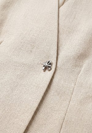 Primo piano di tessuto beige testurizzato con una spilla argentata stilizzata a forma di foglia attaccata vicino a un bordo piegato.