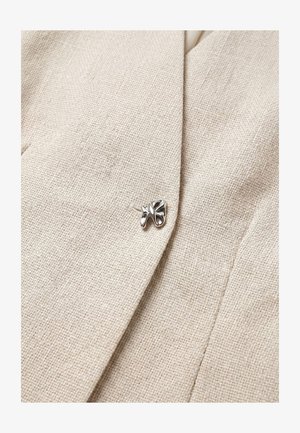 Close-up van beige gestructureerde stof met een zilveren gestileerde bladvormige speld bevestigd nabij een gevouwen rand.