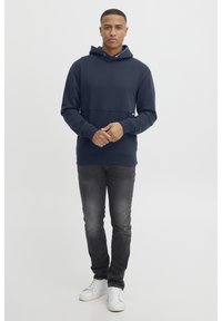 Mornarica kapuca s kengurudžim žepom i rebrastim manžetama, u kombinaciji s tamnosivim trapericama i bijelim tenisicama na neutralnoj pozadini.