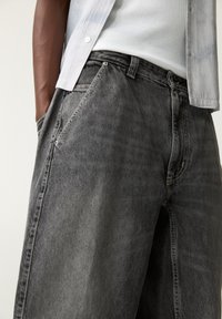 Pantalons en denim gris avec une coupe ample, une taille plate et des poches latérales. Le tissu a une texture délavée avec des détails de surpiqûre contrastants.