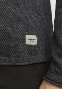 Suéter de punto gris oscuro con tejido texturizado, que muestra una etiqueta cosida con "JACK & JONES EST. 1990" en el dobladillo.