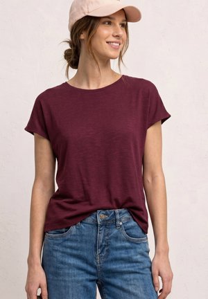 Femme souriante, portant une casquette de baseball rose clair, un t-shirt bordeaux à manches courtes et un jean bleu, debout contre un mur clair uni.