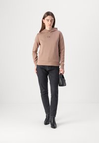 Sudadera con capucha marrón con el logo "CK", acompañada de jeans negros y botines. Diseño minimalista, textura de la tela visible. Bolsa negra en la mano.