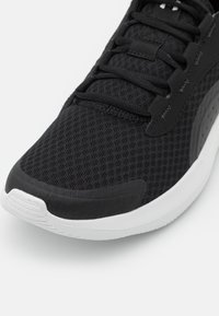 Chaussure de sport noire avec une tige en mesh respirant et des accents synthétiques lisses, dotée d'une semelle en caoutchouc blanche et de lacets noirs plats.