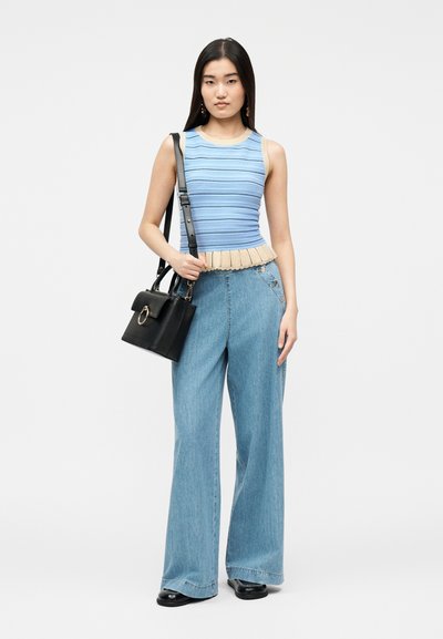Νεαρή γυναίκα όρθια, φοράει αμάνικο πλεκτό τοπ ριγέ σε μπλε και μπεζ αποχρώσεις, τζιν με ψηλή μέση και φαρδιά μπατζάκια, μαύρα παπούτσια και κρατάει μαύρη τσάντα χειρός.
