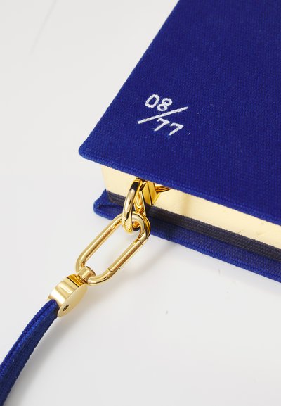 Blue fabric notebook with a textured cover, gold hardware, and a clasp. Features a white embroidered detail "08/77" on the corner.