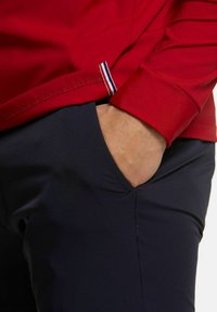 Rotes Langarmshirt mit abgerundetem Saum und einem kleinen gestreiften Akzent. Kombiniert mit navyblauen Hosen, die eine Taschen-Detail aufweisen.