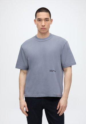 Fiatal férfi laza, rövid ujjú szürke pólót visel elöl "Karl Lagerfeld Jeans" felirattal, egyszínű háttér előtt áll.