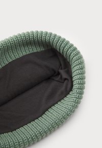 Strikket grønn hue med ribbestrikket kant, utstyrt med et glatt mørkt stofffôr på innsiden. Formen er rund og designet for hodeplagg.