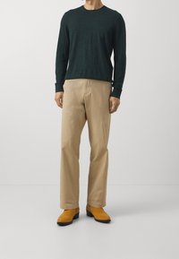 Man som bär en mörkgrön långärmad skjorta, beige byxor och senapsgula mocka ankelstövlar står mot en enfärgad bakgrund.