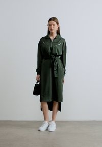 Σκούρο πράσινο φόρεμα πουκάμισο με φερμουάρ στο μπροστινό μέρος, μακριά μανίκια, σφιγμένη μέση και πλαϊνή σχισμή, σε συνδυασμό με λευκά αθλητικά παπούτσια και μικρή μαύρη τσάντα.