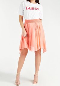 Biała koszulka z czerwonym logo "GUESS" w zestawieniu z koralową, zwiewną spódnicą do kolan z marszczoną talią i koronkowymi detalami.