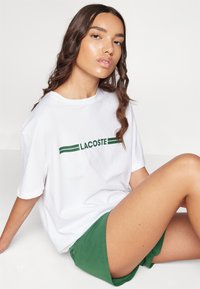 T-shirt en coton blanc avec le texte et des rayures « LACOSTE » en vert. Assorti à un short vert, en position assise contre un fond neutre.