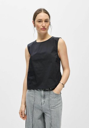 Femme aux cheveux attachés portant un haut noir sans manches et un jean gris clair taille haute, debout avec une main dans la poche devant un fond blanc.