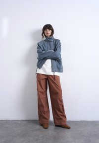 Γυναίκα που στέκεται με σταυρωμένα χέρια, φοράει τζιν μπουφάν με ψηλό γιακά, λευκό πουκάμισο, φαρδιά παντελόνια σε σκουριά χρώμα και καφέ μυτερά παπούτσια σε γκρι δάπεδο.