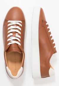 Bruna lädersneakers med vita snören och detaljer, med en vit gummisula och en slät yta. Logotyp på innersulan.
