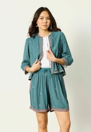 Nő türkizkék hímzett dzsekiben és hozzáillő rövidnadrágban, fehér felsővel, kezével a dzseki elejét igazítva pózol.