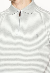 Grå och vitrandig pikétröja med dragkedjekrage och liten broderad logotyp på vänster sida. Tygblandning i bomull med en slät textur.