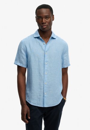 Mężczyzna w jasnoniebieskiej koszuli z krótkim rękawem i kołnierzykiem, połączonej z ciemnymi spodniami i zegarkiem na nadgarstku, na białym tle.