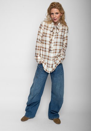 Frau trägt eine weiß-braune karierte Bluse mit Schleife und weite blaue Jeans und steht mit den Händen in den Taschen.