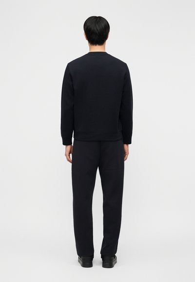 Άνδρας που στέκεται με την πλάτη γυρισμένη, φοράει μαύρο μακρυμάνικο πάνω μέρος, μαύρο παντελόνι και μαύρα παπούτσια, μπροστά σε απλό ανοιχτόχρωμο φόντο.