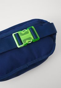 Mørkeblå stofpose med en grøn plastspænde. Har en struktureret overflade og en justerbar rem. Kompakt, rektangulær form.