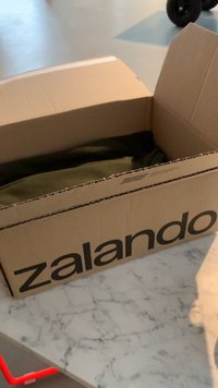 Kartónová krabice s nápisem "zalando" vytištěným tučnými černými písmeny, částečně otevřená, aby odhalila složenou látku olivové barvy uvnitř, na mramorovém povrchu.