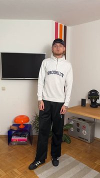 Svetlosivá mikina na zips s nápisom "BROOKLYN", v kombinácii s čiernymi teplákmi. Oblečená s čiernymi teniskami, na ktorých sú viditeľné tetované ruky.