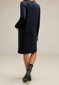  marinblå stickad klänning med långa ärmar, rundad hals och en avslappnad passform. Bärs med mörkgröna ankelboots. Slät textur, minimal detaljering.