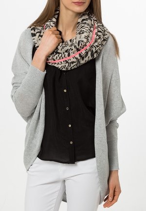 Femme portant une chemise noire à boutons, un cardigan gris clair, un pantalon blanc et une écharpe infinie beige à motifs avec une bordure rose.