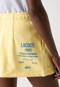 Geltona sportinė sijonas, pasižymintis lygiu tekstūru, su mėlynu logotipo spaudiniu ir tekstu ant nugaros bei Lacoste krokodilo akcentu.