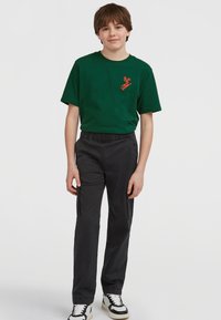 Grön bomullströja med en orange grafisk design, i kombination med svarta byxor med elastisk midja och sidofickor samt svarta och vita sneakers.