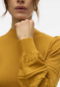 Gul tröja med ribbade hög hals och texturerade spetsärmar. Mjuk tyg, detaljerad med ett blommigt spetsmönster på ärmarna.