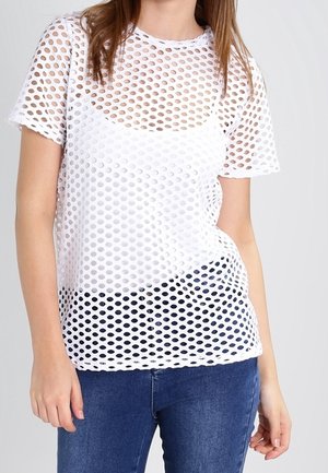 Femme portant un top en mesh blanc à manches courtes sur un débardeur blanc, associé à un jean bleu, debout devant un fond uni.