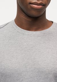 Κοντινό της κάτω πλευράς προσώπου και του λαιμού ενός ατόμου που φοράει ανοιχτογκρι μπλουζάκι με στρογγυλή λαιμόκοψη, σε απλό λευκό φόντο.