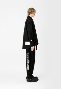 Camisa negra de gran tamaño con tejido texturizado, correas laterales y un bolsillo blanco. Pantalones negros con patrones gráficos y zapatos negros gruesos.