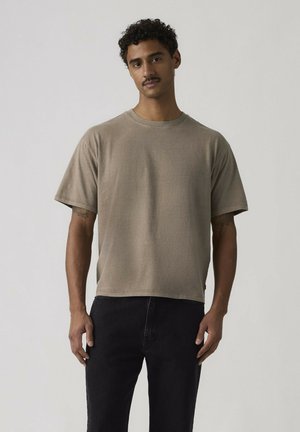 Mand med krøllet hår og overskæg iført en brun t-shirt med korte ærmer og sorte bukser mod en ensfarvet lys baggrund.