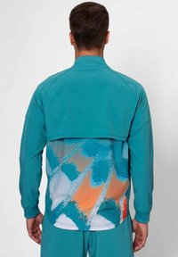 Chaqueta deportiva en color azul verdoso con un diseño en la parte trasera en capas que presenta un gráfico en azul, naranja y blanco. Hecha de un tejido ligero y texturizado.